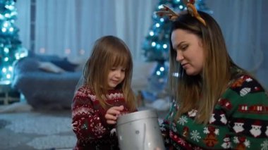 Kız, Noel ağacının yanında annesinin kollarında hediye olan bir kutuyu açıyor. Yeni yıl ve Noel 'in kutlamaları ve duyguları. Mutlu aile gülümsemesi sürprizi. Yüksek kalite 4k görüntü
