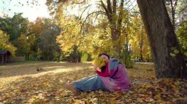 Sonbahar parkında piknikte gülümseyen mutlu kadın. Kız kendini aptal durumuna düşürüyor ve yüzünü bir buket kabarık yaprakla kaplayarak gülümsüyor. Yüksek kalite 4k görüntü