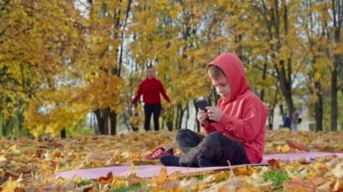 Annesi parkta fitness yaparken, oğlu telefonda oyun oynuyordu. Çocuk çizgi film izliyor, kadın antrenman yapıyor ve spor yapıyor. Yüksek kalite 4k görüntü