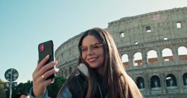 Roma 'daki Colosseum yakınlarında telefonuyla selfie çeken mutlu bir bayan turist. Seyahatlerde hayatın eğlenceli anları, tatilde gülücükler ve neşe. Yüksek kalite 4k görüntü