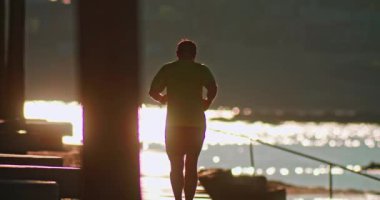 Kulaklıklı bir adam gün batımında deniz kenarında gölgede koşar. Spor ve aktif yaşam tarzı sahilde koşuyor. Yüksek kalite 4k görüntü