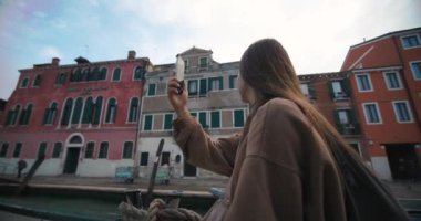 Bir kız, Venedik kanallarında bir teknede yelken açarken fotoğraf çekiyor. Romantizm şehrine güzel mimarisi olan bir turist gezisi, hafta sonu için bir yaşam tarzı tatili. Yüksek kalite 4k görüntü