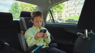 Çocuk telefonda oyunlar oynuyor ve okula arabayla gitti. Akıllı telefonu olan bir çocuk emniyet kemeri takılı bir şekilde çocuk oto koltuğunda oturuyor. Yüksek kalite 4k görüntü