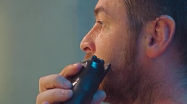 Beyaz erkek sakalını kesme makinesiyle tımar ediyor. Evde temizlik ve yüz bakımına odaklanıyor. Yüksek kalite 4k görüntü