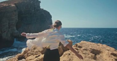 Okyanus kıyısındaki bir uçurumun tepesinde ellerini kaldıran bir kadının yavaş çekim videosu. Seyahat özgürlüğü hissi ve deniz kenarında yürüyüş turizmi. Yüksek kalite 4k görüntü
