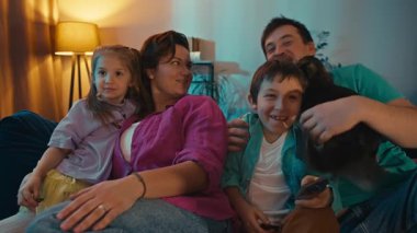TV 'de birlikte film izleyen mutlu bir beyaz aile. Çocuklar ve ebeveynler kanepede oturup televizyon izlerken gülümseyerek tepki veriyorlar. Yüksek kalite 4k görüntü