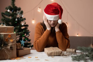 Yılbaşı şapkası takmış yorgun genç bir kadın gözlerini çekti hediye paketi siparişi aldı, arka planda bir Noel ağacı ve ışıklar, yeni yıla hazırlık konsepti, mutlu noeller, 2023