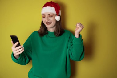 Yeşil kazak ve Noel Baba şapkası giymiş neşeli genç bir kadın elinde bir cep telefonu, yeşil bir arka planda izole edilmiş, başarılı bir jest yapıyor. Mutlu yıllar 2023 tatil konsepti.