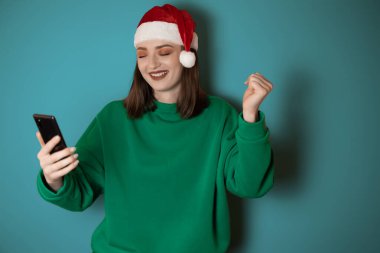 Yeşil kazak ve Noel Baba şapkası giymiş neşeli genç bir kadın elinde bir cep telefonu, yeşil bir arka planda izole edilmiş, başarılı bir jest yapıyor. Mutlu yıllar 2023 tatil konsepti.