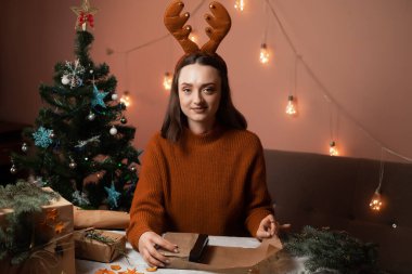 Mutlu bir genç kadın Noel için hazırlanıyor ve hediyeler paketliyor, kızın kafasında yeni yıl geyik boynuzları var, arka planda bir Noel ağacı ve ışıklar, yeni yıla hazırlık konsepti, mutlu noeller, yeni yıl 2023