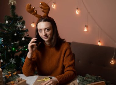 Mutlu bir genç kadın telefonla sipariş verir, kafasında yeni yıl geyik boynuzları vardır, hediye paketi siparişi alır, arka planda bir Noel ağacı ve ışık, yeni yıla hazırlık konsepti, mutlu noeller, yeni yıl 2023