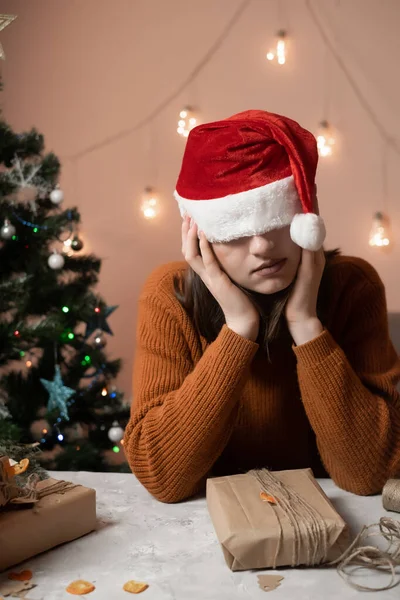 tired young woman in a New Year\'s hat pulled over her eyes taking an order for gift wrapping, in the background a Christmas tree and lights, the concept of preparing for the new year, merry christmas, new year 2023