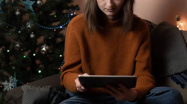 a young woman holds a phone, a tablet in her hand and fulfills an order by phone, taking an order for gift wrapping, in the background a Christmas tree and lights, the concept of preparing for the new year, merry christmas, new year 2023