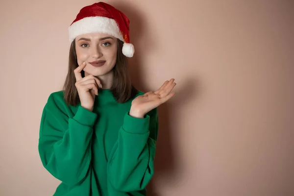 Merry surprised young woman wear xmas sweater Santa hat posing pointing hands arms aside indicate on workspace area isolated on plain pastel light green background. Happy New Year 2023 holiday concept