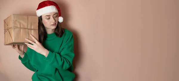 young woman in a green sweater and in a New Year's hat holds a gift box on a simple green background. Holiday concept Happy New Year 2023, chrome key, merry christmas