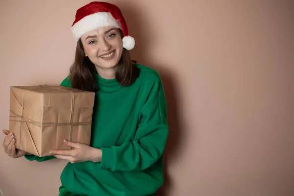 young woman in a green sweater and in a New Year\'s hat holds a gift box on a simple green background. Holiday concept Happy New Year 2023, chrome key, merry christmas