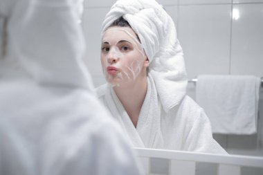 Young caucasian woman in a white bathrobe and with a towel on her head removes a moisturizing sheet mask from her face, looking in the mirror