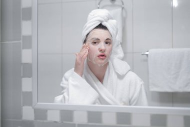 Young caucasian woman in a white bathrobe and with a towel on her head removes a moisturizing sheet mask from her face, looking in the mirror
