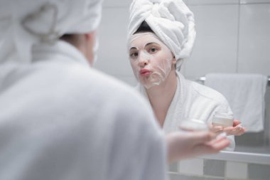 Sabah duştan sonra kafasında havluyla aynaya yansıyan kadın gündüz yüz kremi sürer. Yakın çekim, ilk kırışıklıkları engeller. Cilt bakım kremi, nemlendirici krem, güzellik tedavisi, cilt bakımı gibi konuları önemser.