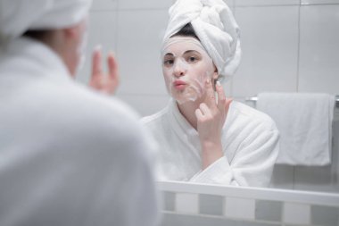 Sabah duştan sonra kafasında havluyla aynaya yansıyan kadın gündüz yüz kremi sürer. Yakın çekim, ilk kırışıklıkları engeller. Cilt bakım kremi, nemlendirici krem, güzellik tedavisi, cilt bakımı gibi konuları önemser.