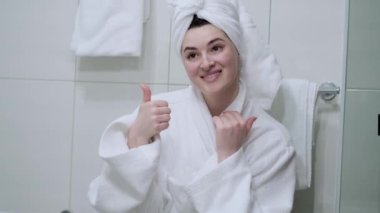 Banyodaki genç bir kızın yakın plan portresi. Başparmaklarını gösteriyor ve kameraya bakıyor. Kafasında beyaz bir bornoz ve beyaz bir havlu var.