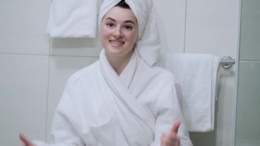 Pürüzsüz Sağlıklı Kadın, Aynaya Bakış, Yüzüne Nazikçe Dokunmak, Gülümsemek. Beyaz bornozlu kız mankeni portresi ve kamera için havlu pozu. Cilt Konsepti.