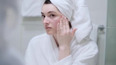 Pürüzsüz Sağlıklı Kadın, Aynaya Bakış, Yüzüne Nazikçe Dokunmak, Gülümsemek. Beyaz bornozlu kız mankeni portresi ve kamera için havlu pozu. Cilt Konsepti.