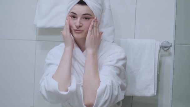 バスローブの女性は鏡を見て顔のマッサージをします 指先で肌に触れ 自宅で簡単に効果的な抗しわ治療をお楽しみください ミームの除去と防止 — ストック動画
