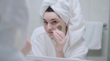 Kız yeşim taşıyla yüz masajı yapıyor, banyoda, kafasında beyaz bir bornozla havluyla dolaşıyor, kız duştan yeni çıktı, evde bakım konsepti...