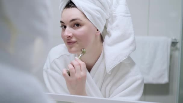 女の子は玉ローラーで顔のマッサージを行い バスルームにあり タオルで彼女の頭の上に白いバスローブを着ている 女の子はちょうどシャワーから出て ホームケアの概念 — ストック動画