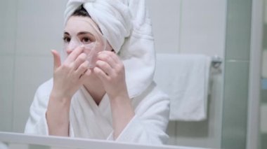 Kozmetik Japon maskesi takan ve banyodaki kameraya bakarken aynaya bakan bir kadın.