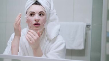Kozmetik Japon maskesi takan ve banyodaki kameraya bakarken aynaya bakan bir kadın.
