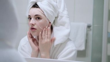 Kozmetik Japon maskesi takan ve banyodaki kameraya bakarken aynaya bakan bir kadın.