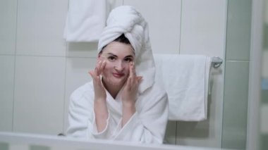Kozmetik Japon maskesi takan ve banyodaki kameraya bakarken aynaya bakan bir kadın.