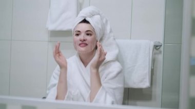 Kozmetik Japon maskesi takan ve banyodaki kameraya bakarken aynaya bakan bir kadın.