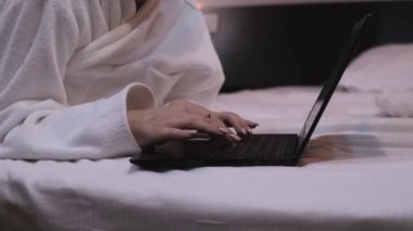 Dizüstü bilgisayarda çalışan kız. Yatak odasındaki beyaz önlüklü kadın yatakta yatıyor ve iki eli dizüstü bilgisayarın klavyesinde. kaybet-up