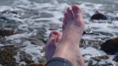 Bir kızın bacaklarının dalga tarafından ezildiği yakın plan bir fotoğraf. Çakıl taşlı deniz kıyısı gürültülü deniz dalgaları dişi bacakları üzerinde