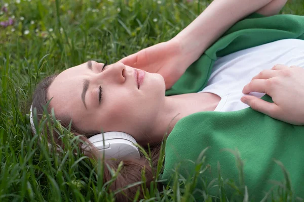 A teenage girl listens to music on headphones, lies on the green grass in the park, smiles and shows her tongue in a white t-shirt and a green sweater thrown over, summer mood as a lifestyle