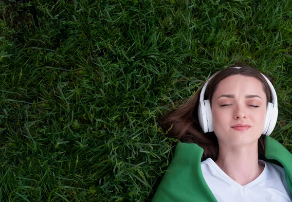 A teenage girl lies on the grass in white headphones, her eyes are closed, listening to music, taking a break from the hustle and bustle, dressed in a white T-shirt, a green jacket is thrown over