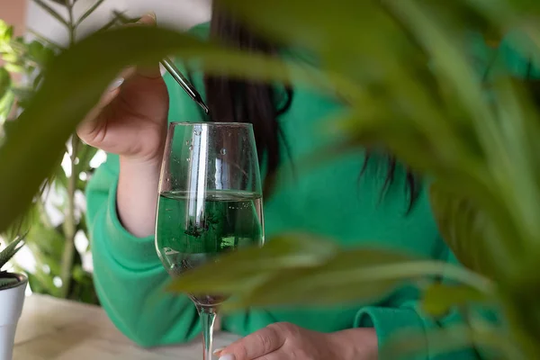 緑のセーターを着た女性がヤシの木の横に立つ水のガラスにクロロフィルの滴を加えます — ストック写真