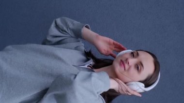 Dikey video, neşeli, kulaklıkla mp3 müzik dinleyen, beyaz ceket, yeşil ceket ve siyah pantolon giyen genç bir kadın tepeden aşağı atılır, müzik eşliğinde dans eder ve şarkı söyler, gri bir sırt üstü.