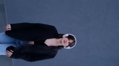 Mutlu bir kızın dans ettiği, kulaklıkla ses dinlediği, kamera önünde mp3 müzik ve dans hareketleriyle eğlendiği dikey video görüntüsü. Neşeli genç bir kadın Gri arka planda şarkılar söylüyor