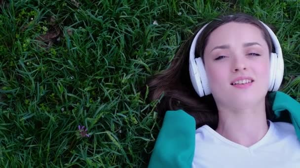 一个松懈的女人躺在草地上 用耳机在公园里听音乐 — 图库视频影像