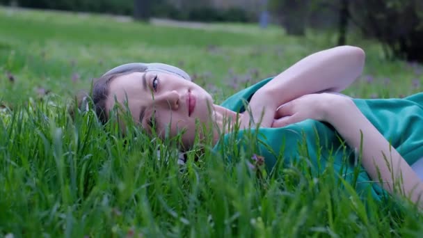 一个松懈的女人躺在草地上 用耳机在公园里听音乐 — 图库视频影像