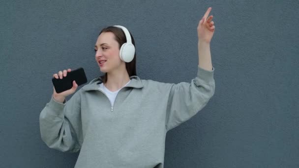 穿着灰色T恤的快乐快乐的年轻高加索女人 带着耳机听音乐 拿着手机 寻欢作乐 在灰墙后背享受假期 — 图库视频影像