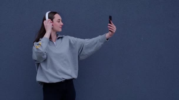 在灰色背景下的手机上带着自拍的自信微笑的女孩 博客写手的生活风格 穿着灰色衬衫 身穿黑色细长的垂直视频 — 图库视频影像