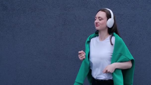 穿着白色T恤和黑色裤子 头戴绿色毛衣的年轻女子戴着耳机跳舞的肖像 带着滑稽的舞步 生活方式和快乐的概念享受着放松的乐趣 — 图库视频影像
