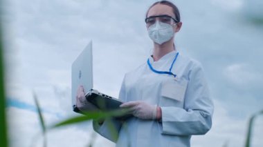 Koruyucu giysili bir araştırma doktoru dizüstü bilgisayarını elinde tutuyor, bitkiler üzerinde bir deney yapıyor, bir laboratuvar çalışanı yeşil bir buğday tarlasında duruyor ve bir filizlenme çiftliğine bakıyor.