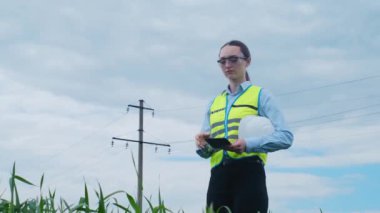 Kadın bir mühendis yüksek voltajlı bir elektrik hattının önünde durur, kaskını çıkarır ve uzaklığa bakar..