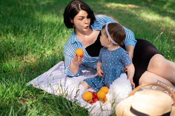 Güzel anne ve tatlı kızı parkta çilek ve mandalina yiyorlar ve piknik günlerinin, aile hafta sonlarının, park tatillerinin tadını çıkarıyorlar.